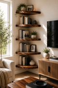 A set of five rustic, live-edge wooden floating shelves mounted on a cream-colored wall in a cozy living room corner. The shelves are styled with books, small potted plants, and framed art, positioned next to a window, a beige sofa, and a wooden TV stand.